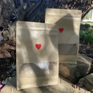 Two kraft paper pouches of Sacred Superfood Shake with red heart stickers, displayed outdoors on natural stone with sunlight and greenery in the background.