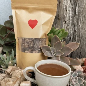 A kraft paper pouch filled with chopped ceremonial cacao sits among succulents and natural stones, with a red heart printed on the pouch. In front of it is a delicate ceramic cup filled with rich, dark cacao drink, set against a weathered wooden backdrop in an outdoor garden setting