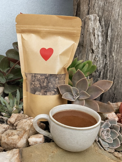 A kraft paper pouch filled with chopped ceremonial cacao sits among succulents and natural stones, with a red heart printed on the pouch. In front of it is a delicate ceramic cup filled with rich, dark cacao drink, set against a weathered wooden backdrop in an outdoor garden setting