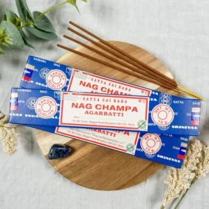 Three blue-and-white Satya Sai Baba Nag Champa incense boxes arranged on a round wooden board, with several brown hand-rolled sticks fanned out on top; soft botanical accents, a navy crystal, and light fabric backdrop create a calm, natural setting.