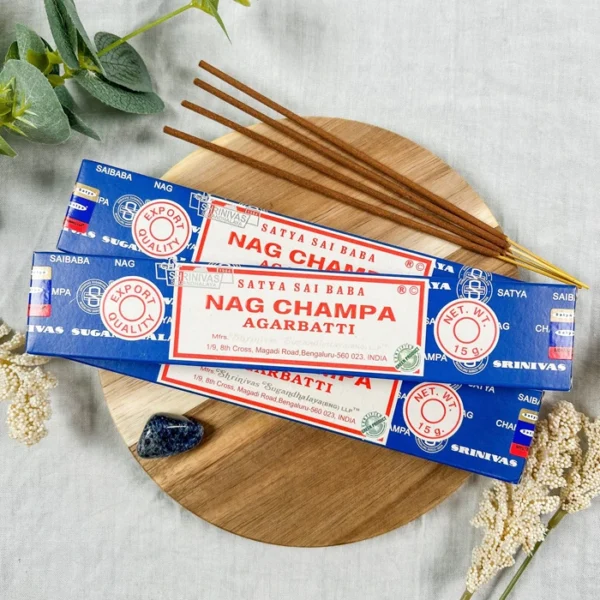Three blue-and-white Satya Sai Baba Nag Champa incense boxes arranged on a round wooden board, with several brown hand-rolled sticks fanned out on top; soft botanical accents, a navy crystal, and light fabric backdrop create a calm, natural setting.